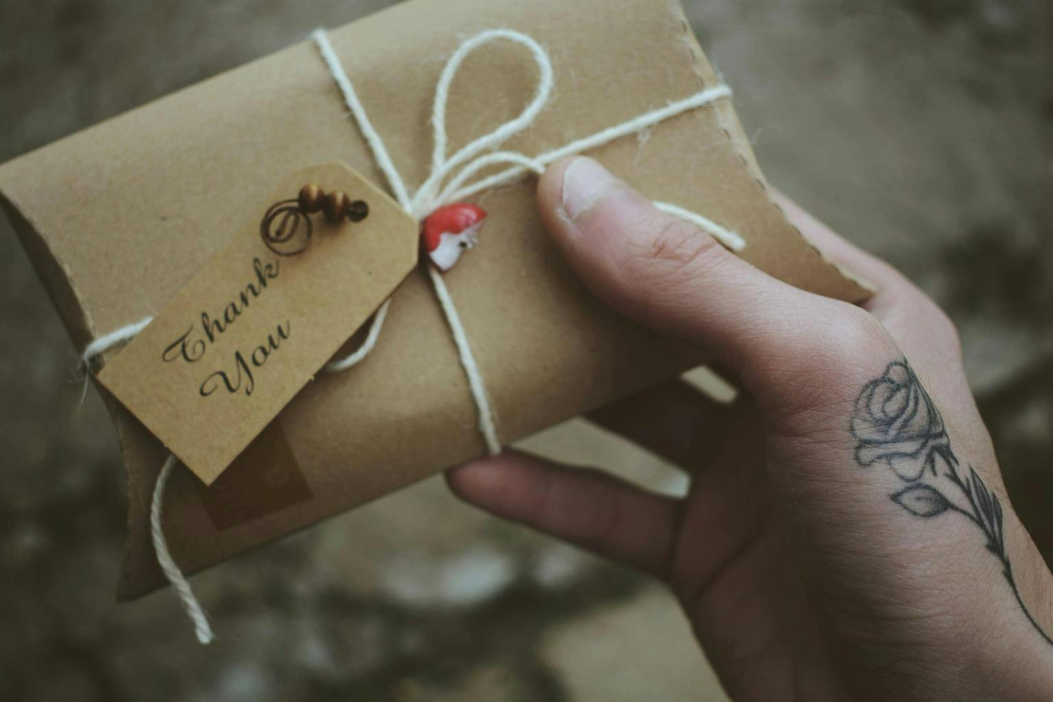 A hand holds a small brown cardboard parcel wrapped with string and small decorations, bearing a note which reads 'Thank you'