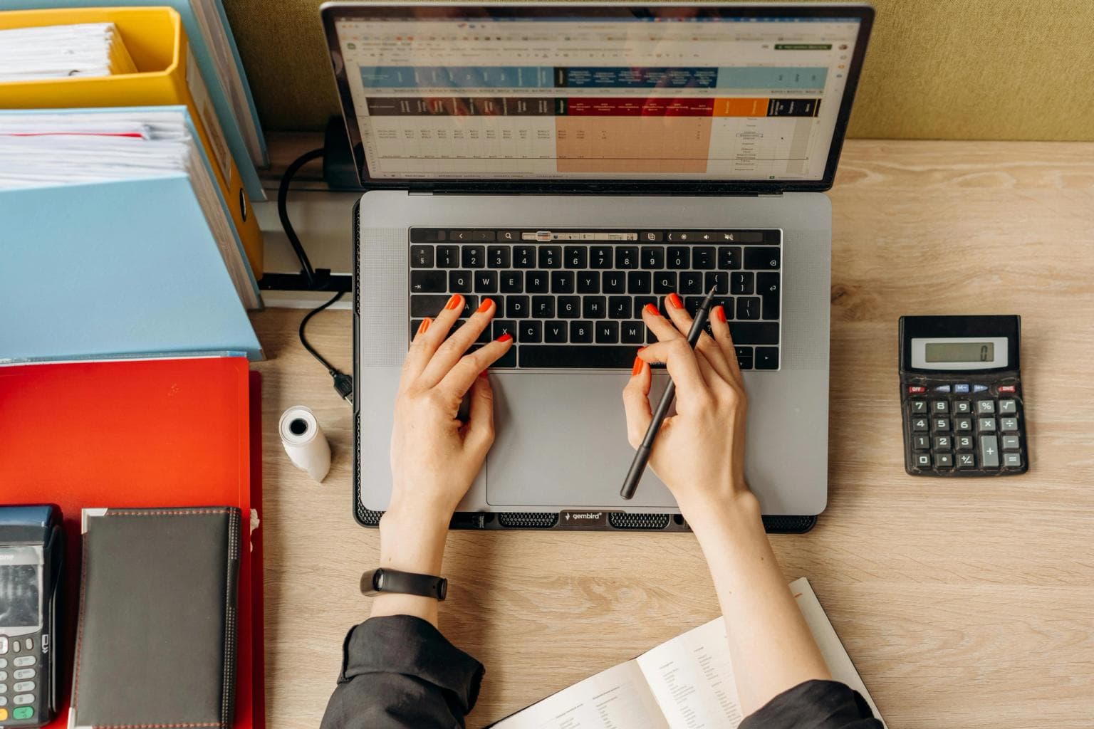 A woman's hands with red nails work at a laptop. A spreadsheet is on screen and a calculator is next to her.