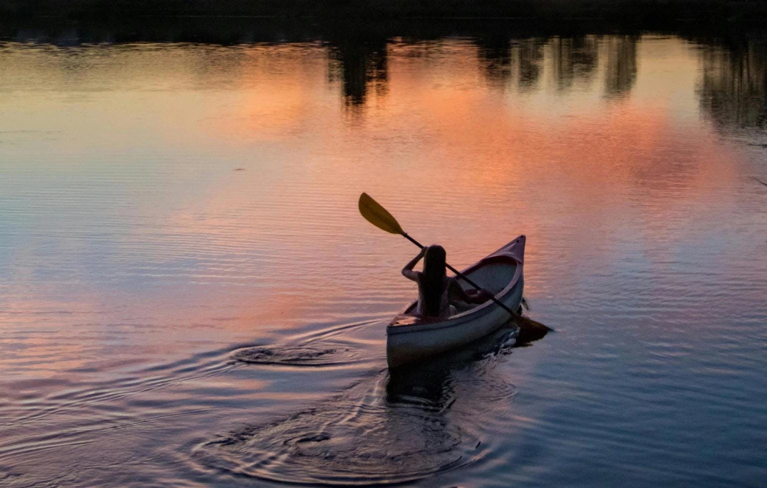 A person in a canoe sits on a lake with sunset colors reflected off the water