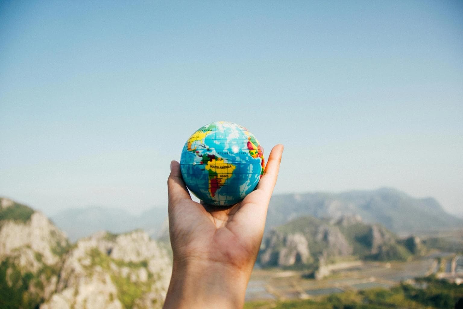 A hand holds up a small globe with mountains in the distance.