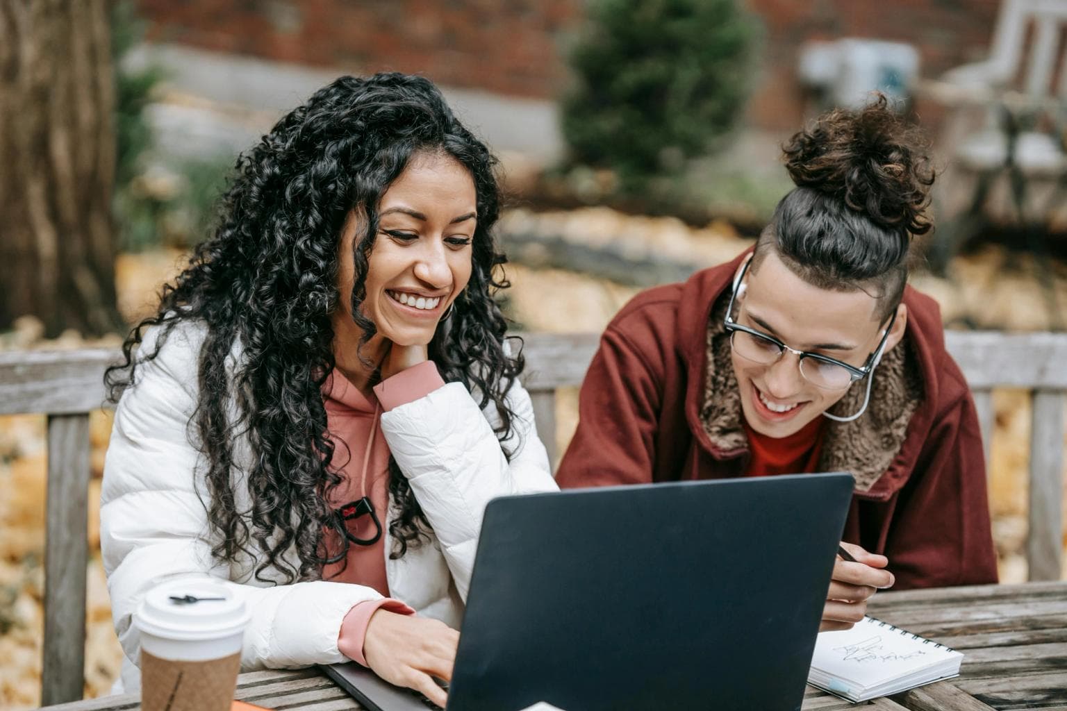 Two women sit outdoors with coffee looking at a laptop together and smiling.