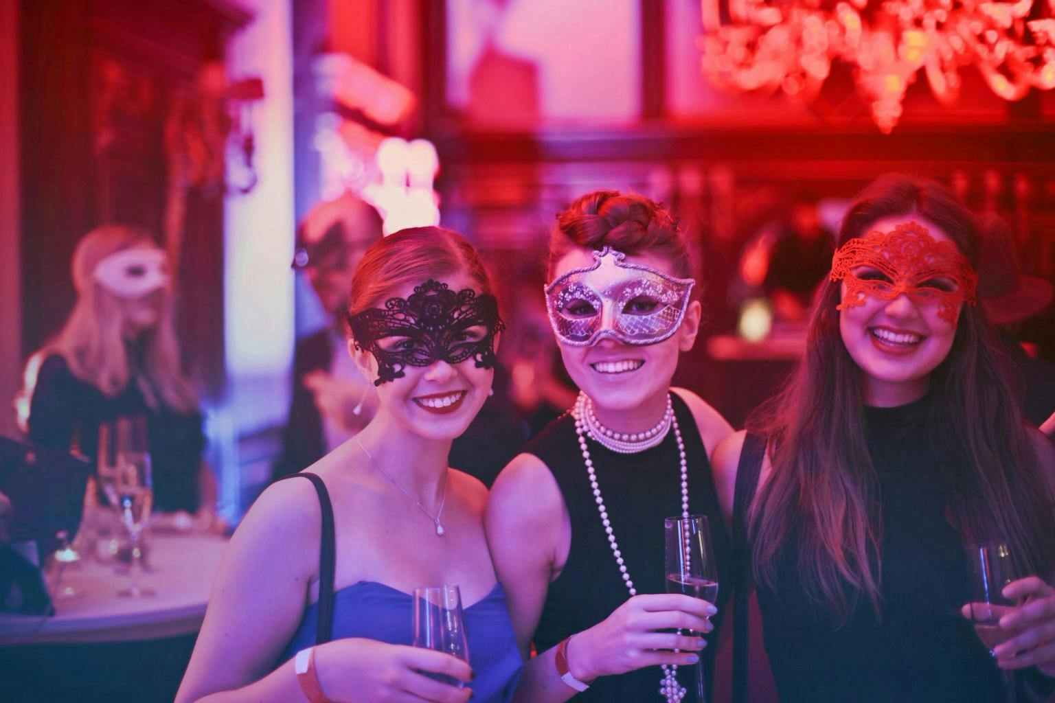 A group of 3 women face the camera smiling and wearing Venetian masks