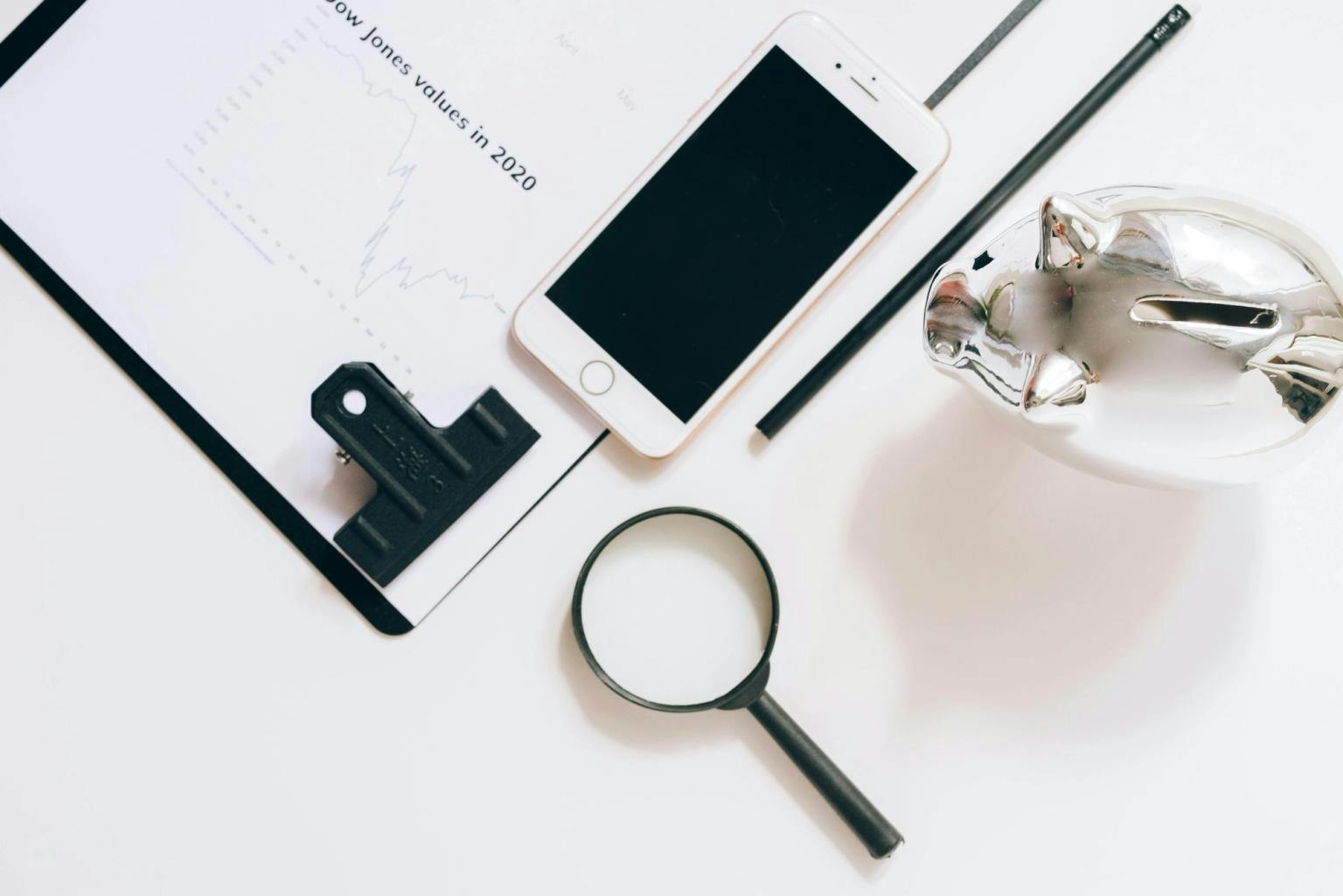 A silver piggy bank sits next to a phone, a clipboard and a magnifying glass