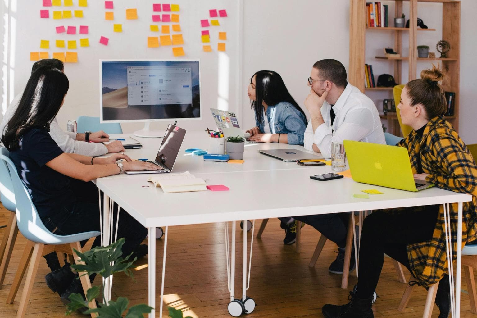 A group of people sit at a desk together discussing what's on screen. A whiteboard with post-it notes is in the background.