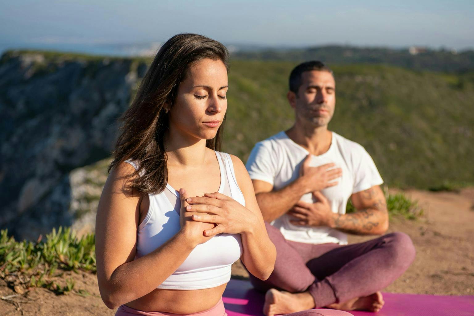 Man and woman sit cross legged on yoga matts with hands on their hearts in a meditative fashion.