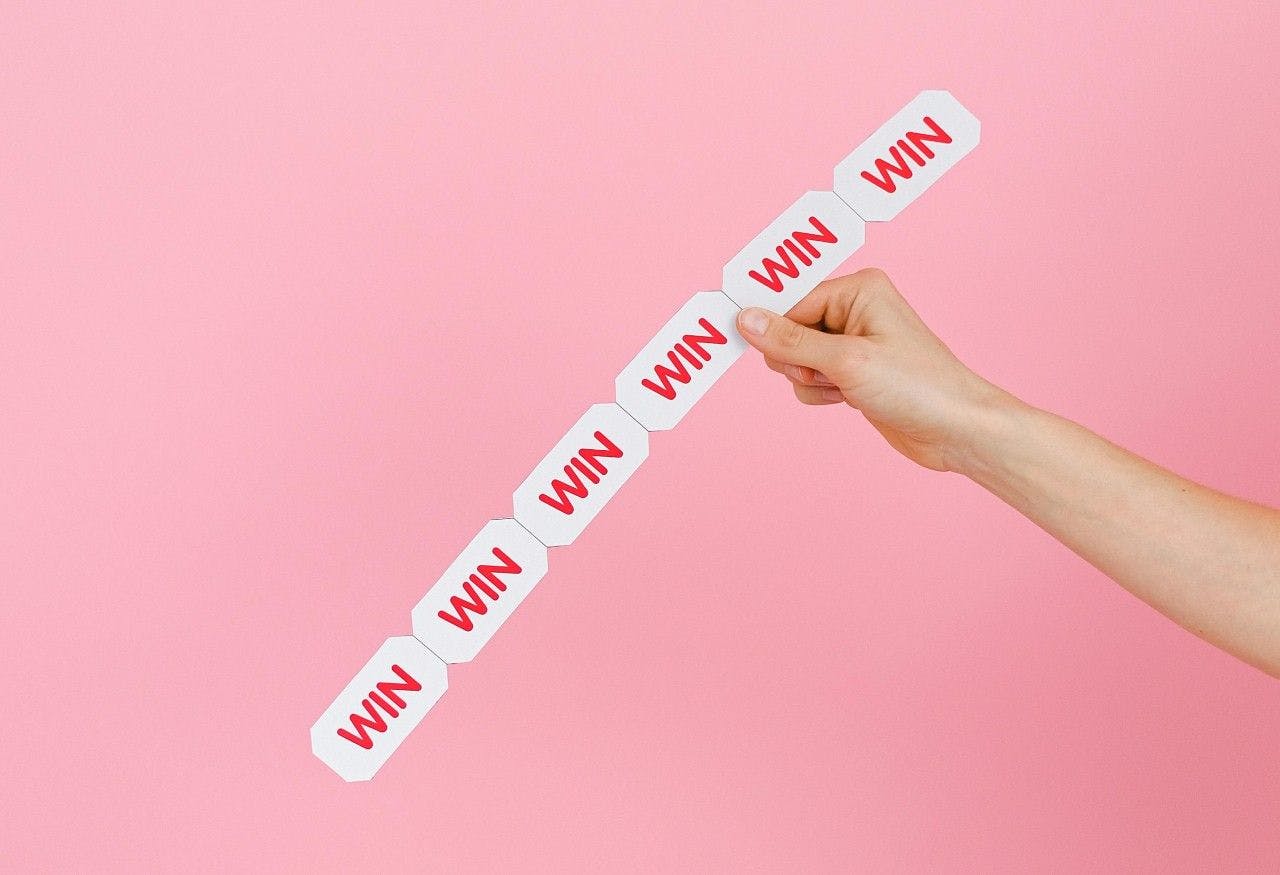 A stream of tickets with the word 'WIN' on them are held up by a hand against a pink backgroud.