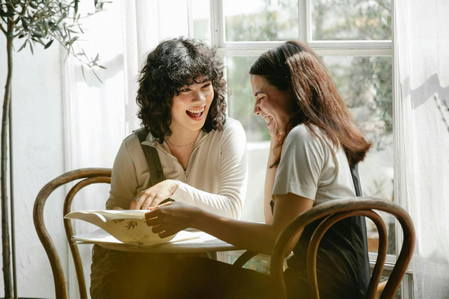Two women sit at a table in a window looking through a document and smiling together