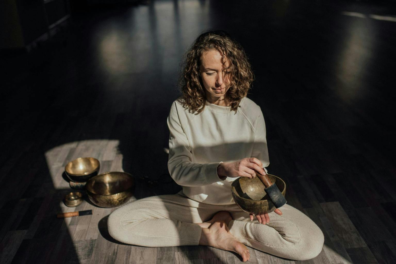 A woman sites with singing bowls, playing one and sitting cross legged in a shady room with light shining on her