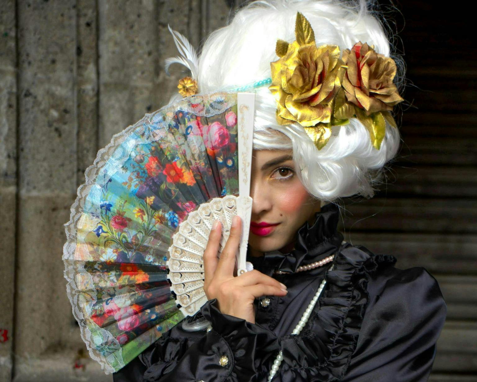 A young woman wearing a romantic period wig with god roses in it, holds a flower painted fan in front of her face