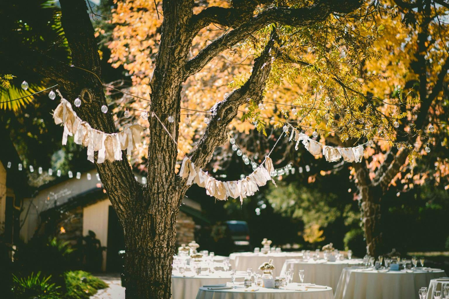 Bunting and lights hangg from trees with round tables set with white cloths underneath