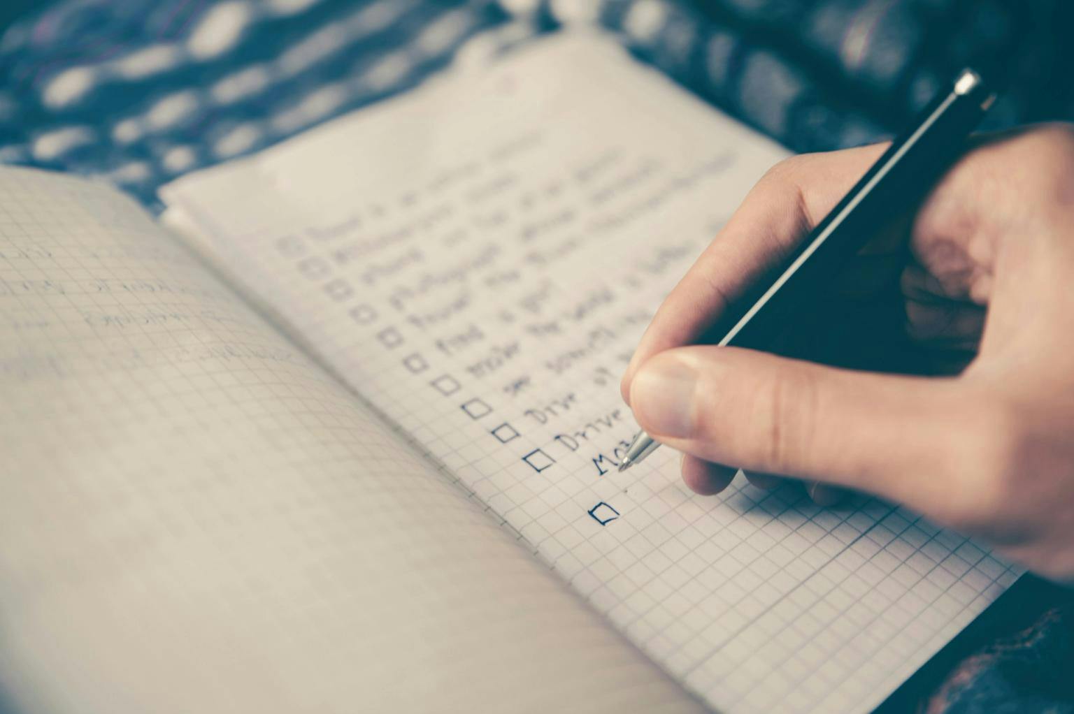 A person's hand holds a pen and is writing into a checklist notebook.