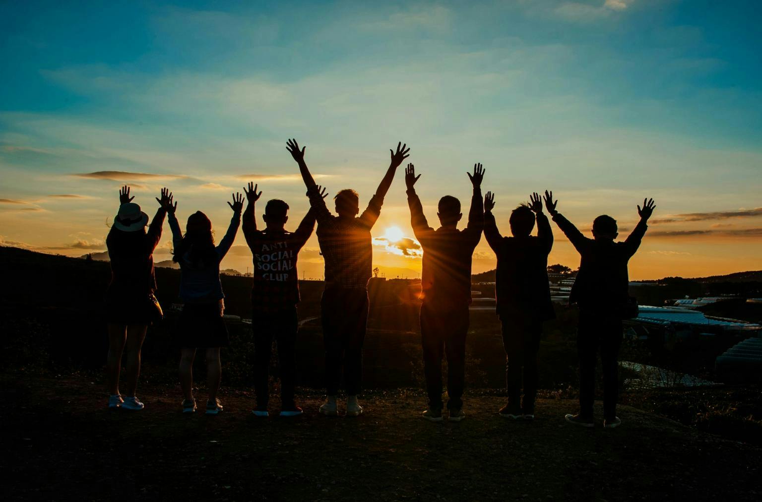 Silhouettes of a group of people watching the sun go down in the distance, arms raised in celebration.