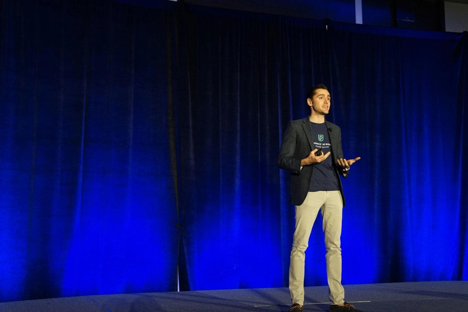 A speaker stands on stage with blue lit curtains behind him.