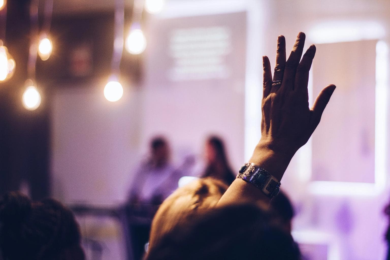 Silhouette of a raised hand in the audience, facing 2 people on stage.