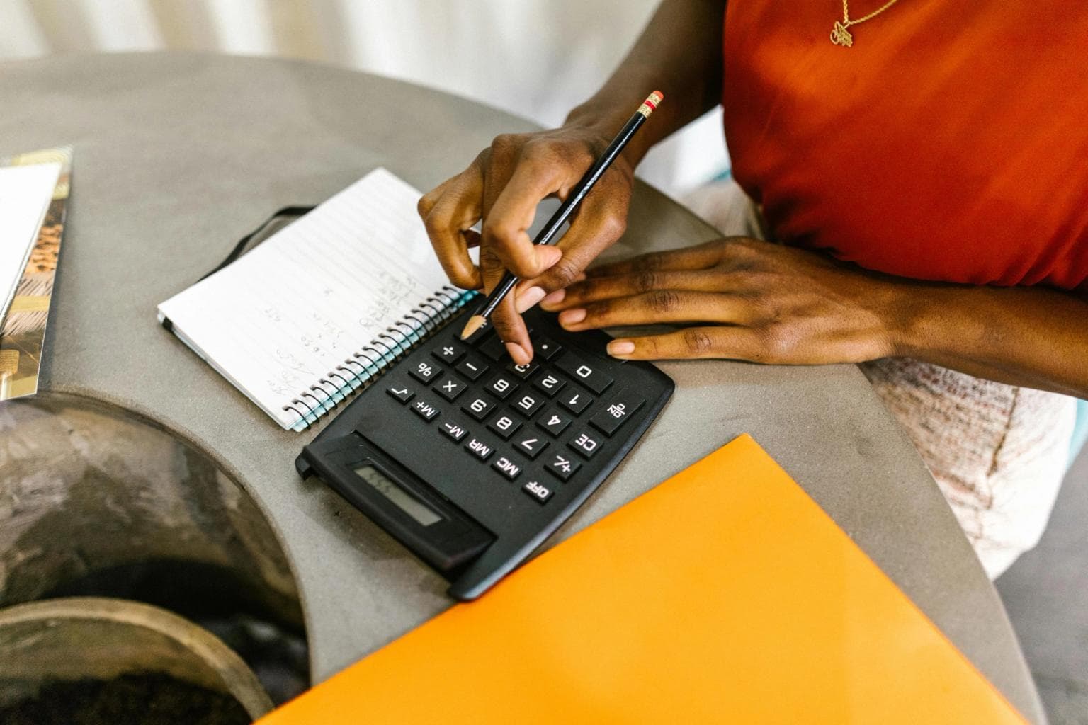 A woman's hands type into a calculator. She holds a pencil and there is a folder on the desk with her.