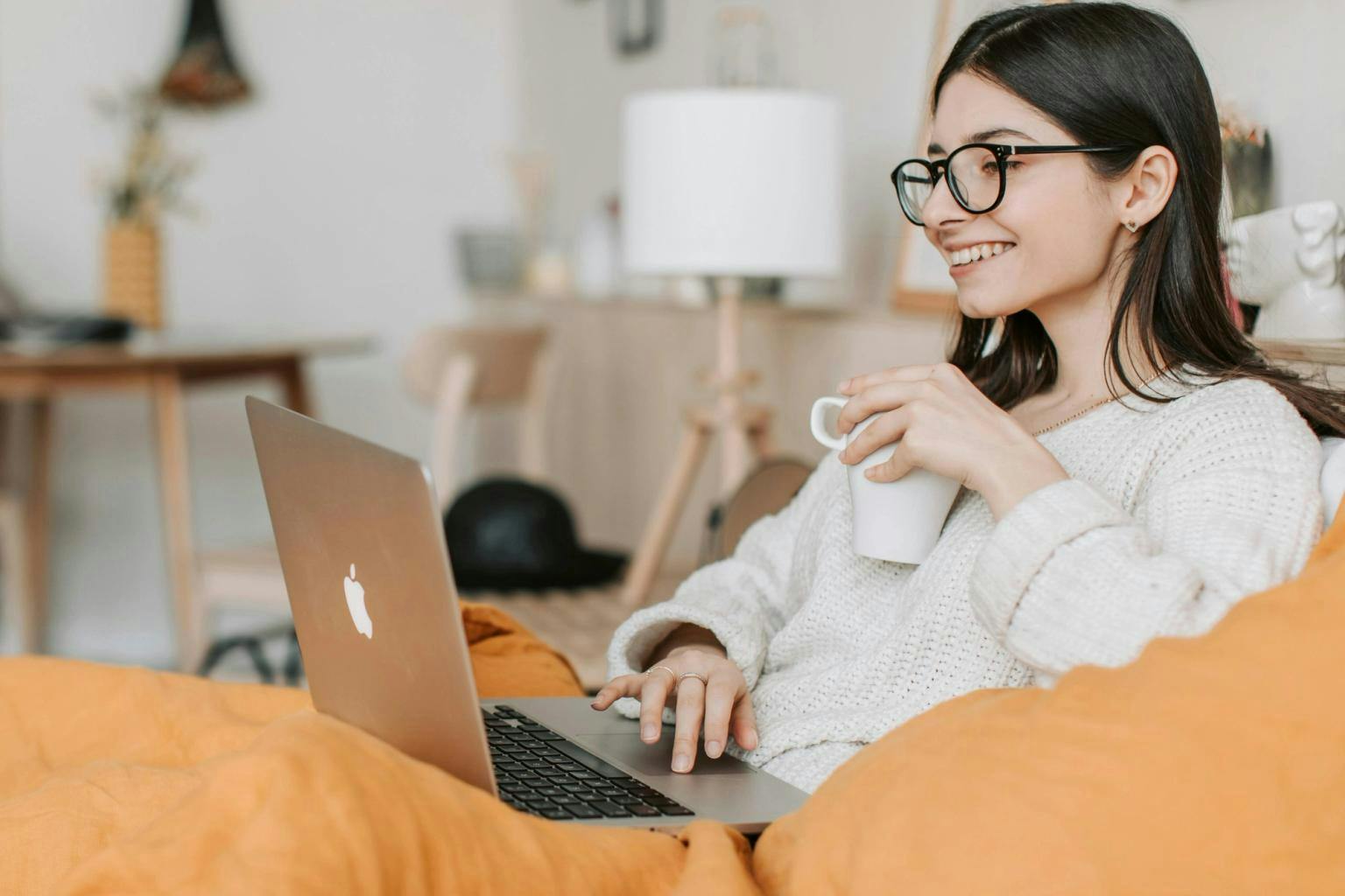 A woman sits at her laptop on a sofa drinking a hot drink and smiling to herself.