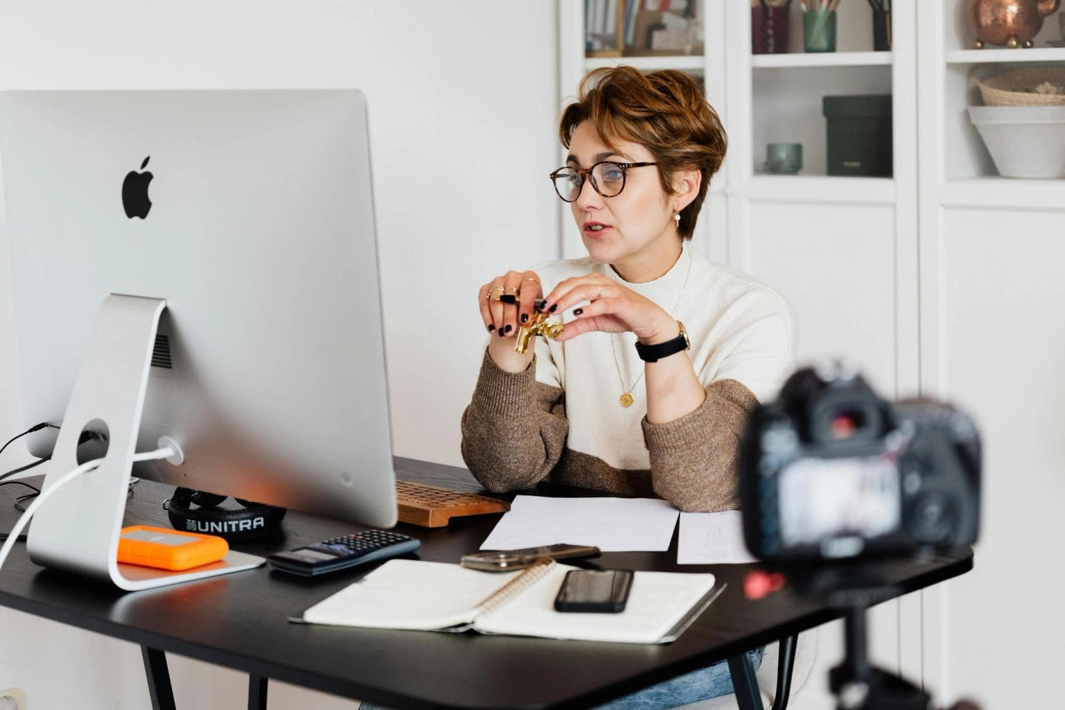 A lady sits at a desk, talking to the screen and a camera that is recording her is in shot