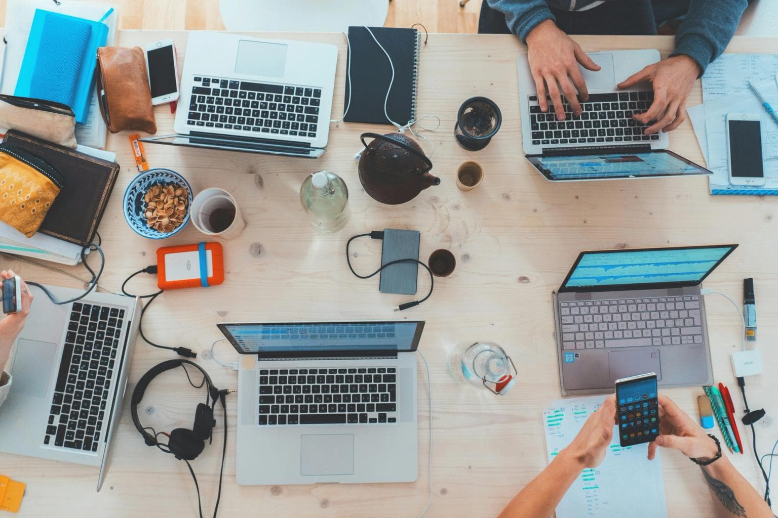 Birds eye view down on a desk with a number of laptops, pads, teas and other gadgets, with some hands in view on the devices.