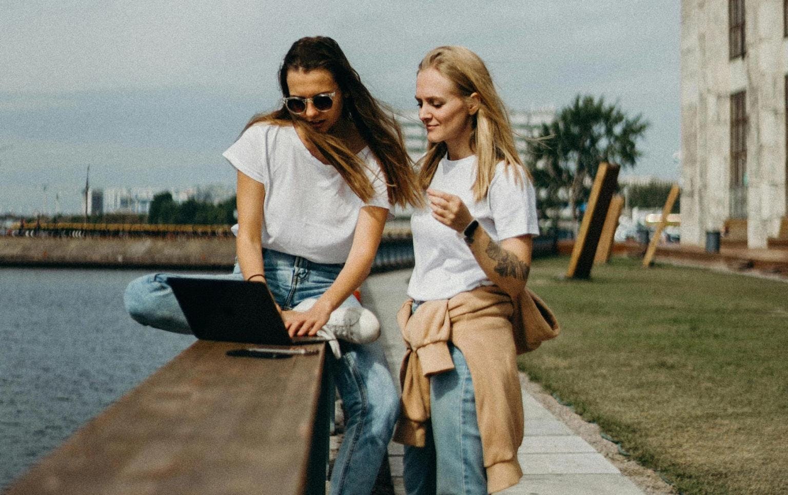 Two women sit outside in a garden overlooking water, working on a laptop 