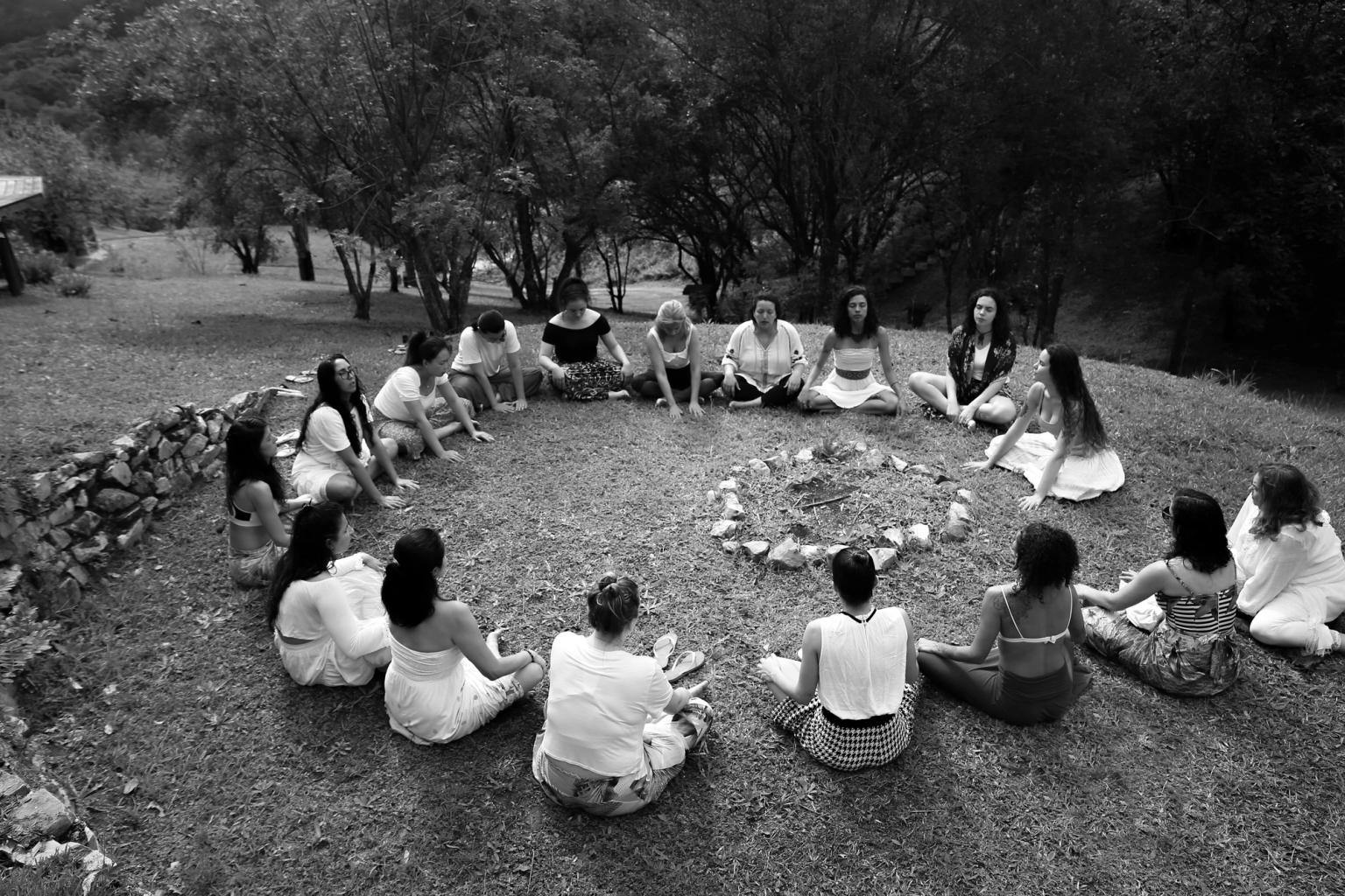 Black and white image of a group of people sat in a large circle outside 