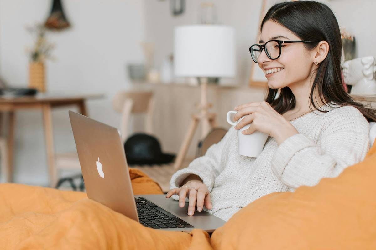 Woman sits on sofa using her laptop drinking a coffee and with a smile on her face.