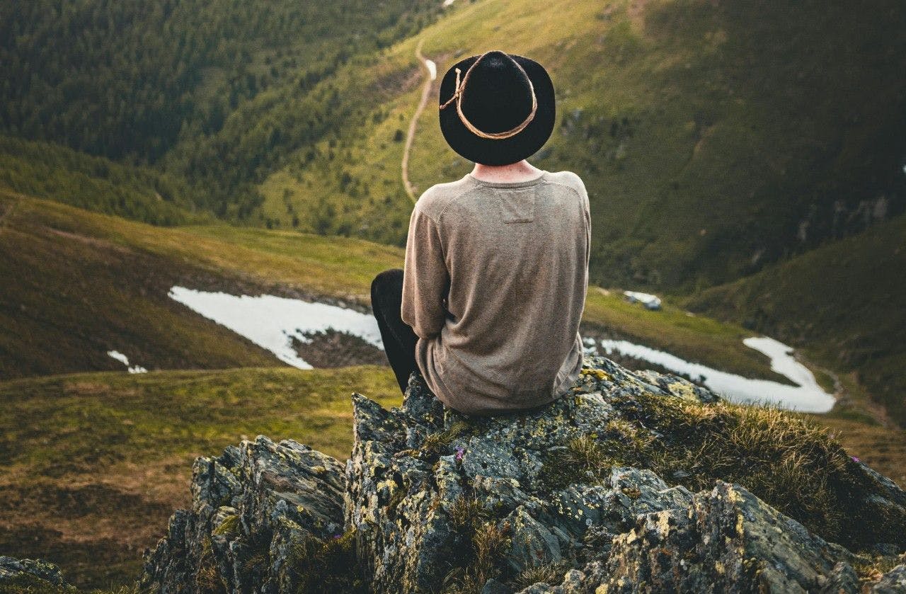 A person wearing a hat sits with their back to the camera on a rock, overlooking a valley below.