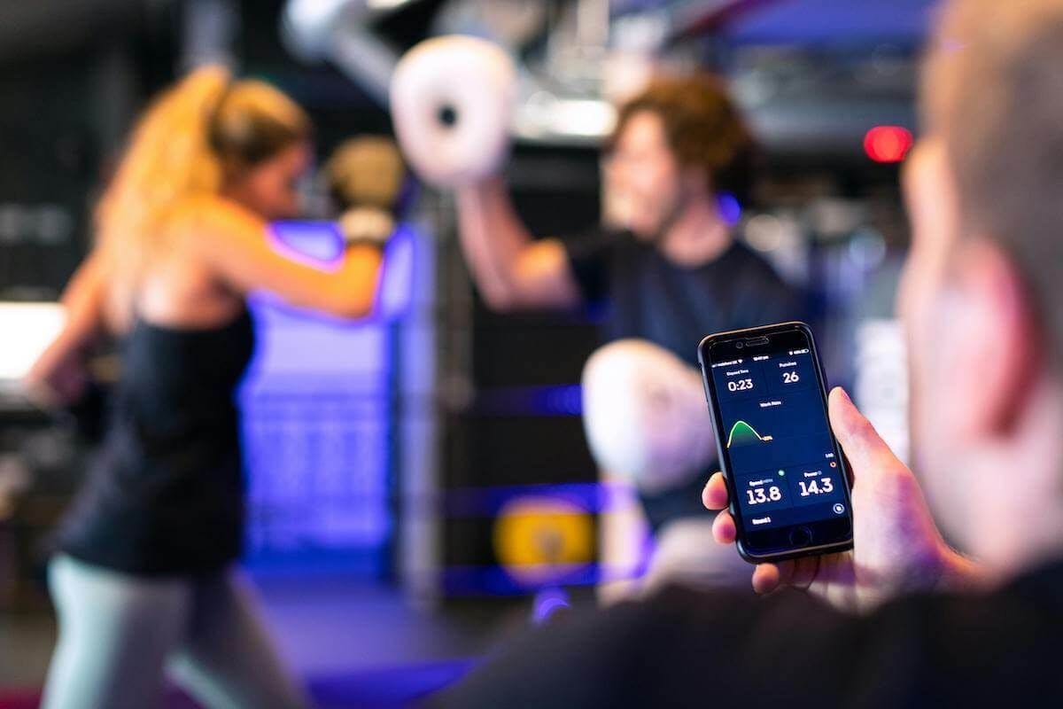 Two women are sparring in the background and a person hold a phone with a fitness app on screen in the foreground